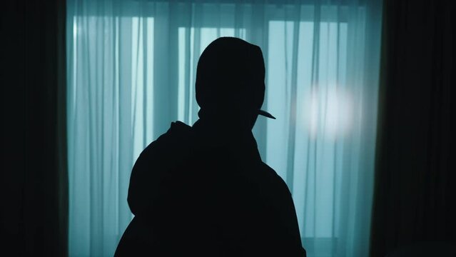A thief looks around in a dark apartment using a flashlight close up. A robber in a balaclava with a backpack and with a crowbar entered the house. The alarm is triggered, the blue red light flashes.