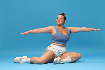 Fototapeta premium Self-care and well being. Young overweight woman stretching on floor in sportswear against blue studio background. Motivation.