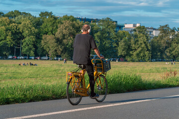 Obraz premium Yellow bike in the park: a man in headphones pedals a city bike with a basket on a sunny day. He enjoys the music, the fresh air and the green scenery of the park with trees and people.