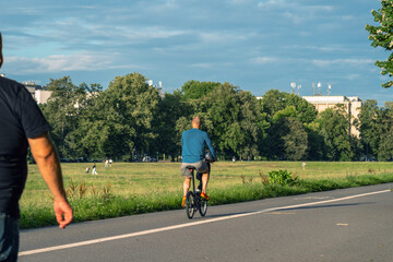 Obraz premium A man in a blue shirt and black shorts rides a black bicycle along a path surrounded by green trees and grass, past people walking in the park. In the background is a white building and a blue sky