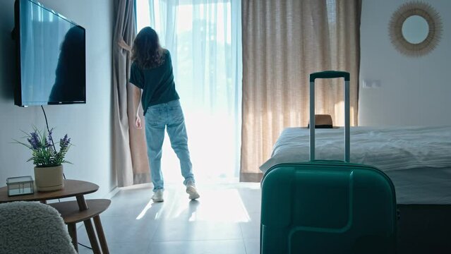 Satisfied joyful woman traveler opens the curtains and sits on the bed in the hotel bedroom after check-in and rejoicing travel start. Enjoy the holiday and vacation. Happy moment