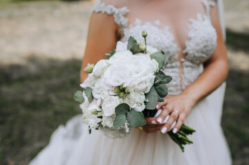 The bride in a white dress holds a bouquet with flowers, roses in her hands outdoors. Wedding portrait, close-up photography.
