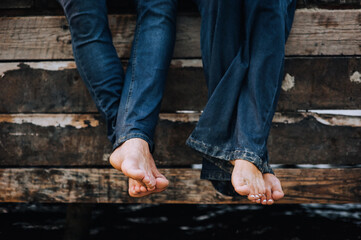 Legs close-up of a barefoot man and woman in jeans sitting on a wooden pier in summer. Close-up photography, portrait.