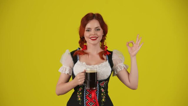 Medium yellow background isolated video of a young German woman, waitress, wearing a traditional costume, holding a glass of dark beer, pointing at it, showing OK sign.