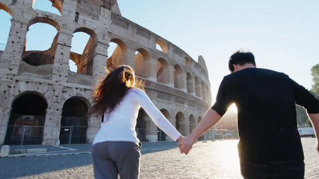 Rear view young adult couple tourist holding hands running together at Colosseum with morning sunrise. happiness people lifestyle, relationship, romantic. Travel destination landmark in Rome, Italy.