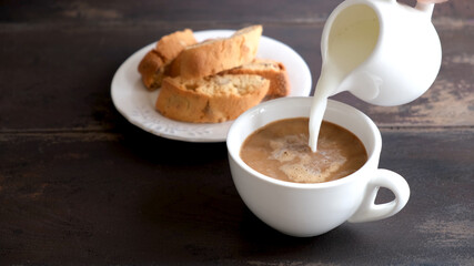 Adding milk cream into black coffee. Coffee with milk and italian cookies Cantucci. Wooden table background