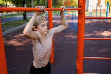 Fototapeta premium A teenager with athletic muscle development holds on to a bar. Street workout on a horizontal bar in the school park.