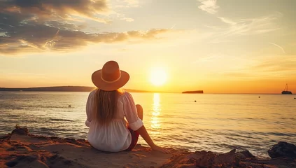 Tuinposter Young woman in hat sitting on the beach and looking at the sunset © Meow Creations