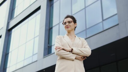 Bottom view young business woman in elegant suit standing outdoor near office building with folded on chest hands, successful and confident head editor of famous magazine
