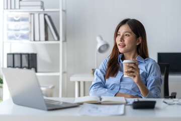 Attractive Asian businesswoman holding hot drink paper cup with smile Relax while working happily with your laptop computer in the office.
