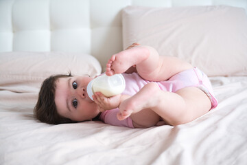 the baby drinks milk from a bottle lying on bed.