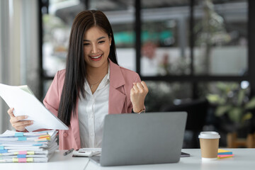 Cheerful and happy businesswoman working on laptop and financial documents in office, attractive smiling female office worker successful.