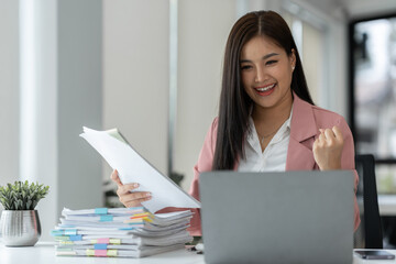 Cheerful and happy businesswoman working on laptop and financial documents in office, attractive smiling female office worker successful.