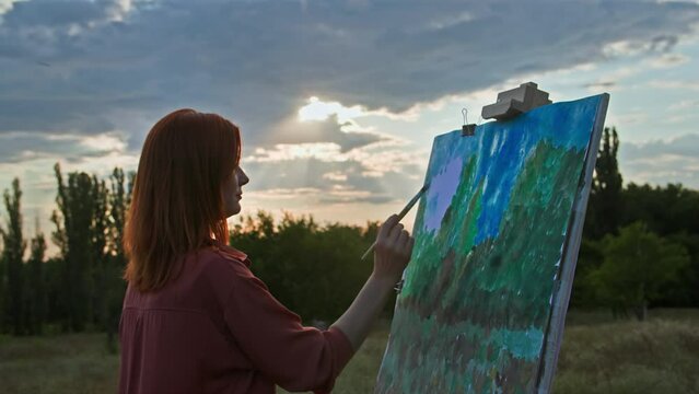 art therapy, charming woman with paint and brush draws a picture using an easel standing in park on background of sun rays