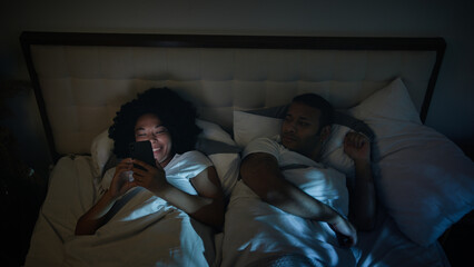 Fototapeta na wymiar African American couple family in bed bedroom night angry frustrated man husband trying sleep offended on wife woman laughing loud using gadget addict disturb sleeping mobile phone addiction problem