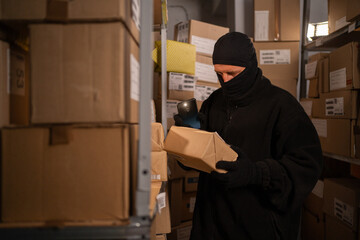 Male thief in black mask steals a parcel in a warehouse at night lightning with flashlight. Security problems in warehouses