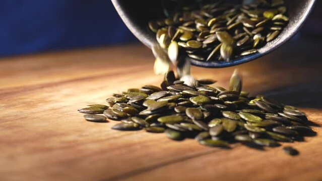 Pumpkin seeds from a ceramic bowl trop out on a wooden board. Close-up.
