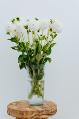 Bouquet of white dahlia flowers in glass vase on an wood vintage stool against the white wall