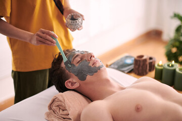 Young female beautician in yellow uniform standing by head of relaxed male client lying on couch...