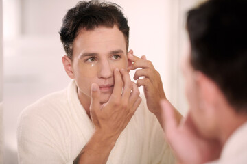 Young serious multiethnic man applying hydrating cream on under-eye area while standing in front of mirror and looking at his face