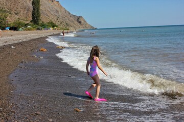 A girl in a bathing suit and pink flippers walks into the advancing foamy wave of the sea on a bright sunny day © Rystemys