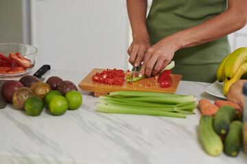 Hands of young woman cutting fresh celery and capsicum on wooden board while standing by kitchen table and cooking breakfast