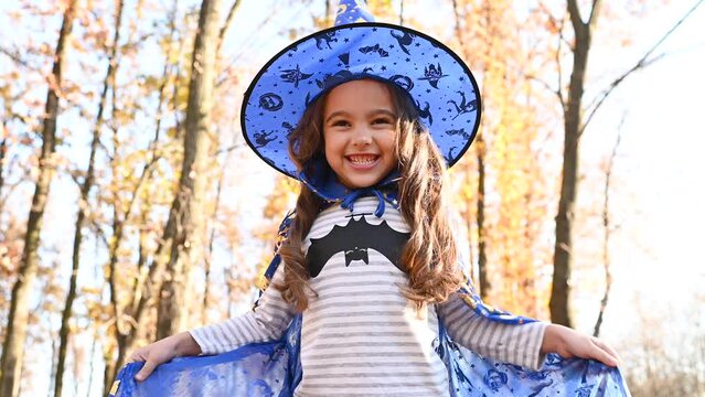 little girl in a Halloween costume and a witch hat scares people and has fun on the background of an autumn forest. Halloween celebration.