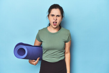 Sporty woman with yoga mat on blue screaming very angry and aggressive.