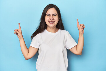 Young Caucasian woman on blue backdrop indicates with both fore fingers up showing a blank space.