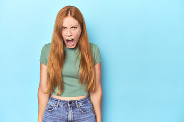Redhead young woman on blue background shouting very angry, rage concept, frustrated.