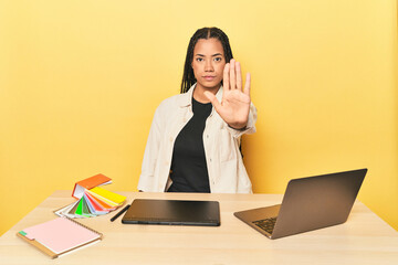 Indonesian designer with tools at table on yellow standing with outstretched hand showing stop...