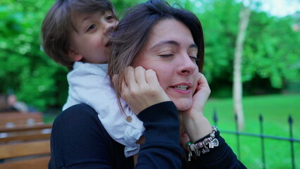 Motherhood moment of little boy hanging on mom's back in open air outside, kissing and hugging mom in authentic love and affection. Caring small boy embraces and kisses parent