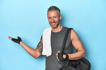 Athlete with gym backpack on blue background showing a copy space on a palm and holding another...