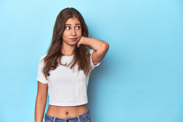 Teen girl in white T-shirt on a blue background touching back of head, thinking and making a choice.