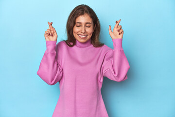 Teen girl in cozy pink sweatshirt on blue crossing fingers for having luck