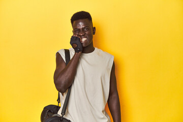 Fit African man with gym backpack on yellow studio background, biting fingernails, nervous and very...
