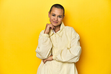 Young woman in jacket, yellow studio backdrop smiling happy and confident, touching chin with hand.