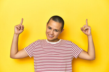 Shaved head woman in red striped tee, yellow backdrop indicates with both fore fingers up showing a...