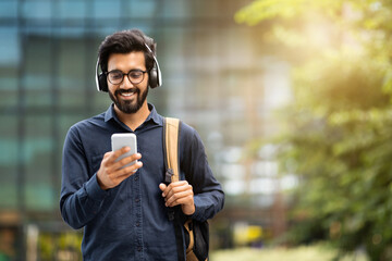 Smiling young indian man wearing headphones holding phone while walking