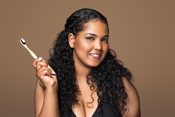 Portrait of beautiful brazilian chubby woman with radiant smile holding wooden toothbrush with paste, brown background