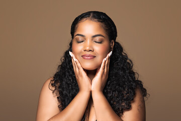 Perfect skin. Beautiful brazilian plus size woman touching her face with pleased face expression, brown background - obrazy, fototapety, plakaty