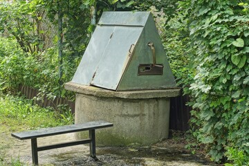 public indoor with concrete circles old with clean water traditional with a gray metal lid a well with a bench stands on the ground in the afternoon near green bushes in the summer on the street