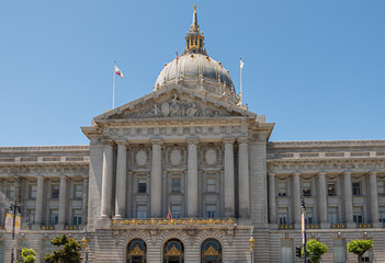 San Francisco, CA, USA - July 12, 2023: Gray stone City Hall east facade pediment above entrance with giant dome under blue sky. Golden decorations.  3 Flags on top: country, state, city