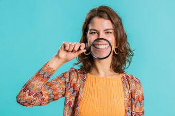 Young woman holding magnifier glass loupe on healthy white teeth, looking at camera with happy expression, showing funny silly face smiling mouth. Brunette girl isolated on blue studio background