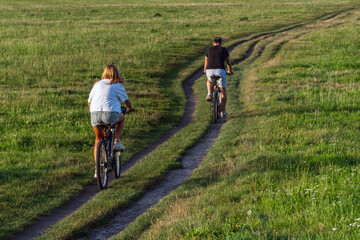 Naklejka premium Two cyclists, a girl and a boy, are enjoying a ride on a rural dirt road. They wear casual clothes and are surrounded by green fields and trees. The picture captures a moment of leisure and relaxation