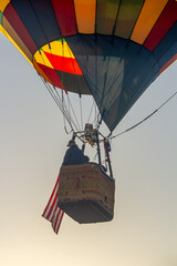 A hot air balloon is ascending into the blue sky. The basket is prominently displayed with an American flag hanging from it. The balloon is yellow, green, blue and red. 