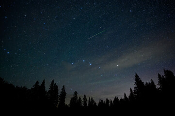 The night sky filled with stars including the Big Dipper and a bright green meteor above a skyline...
