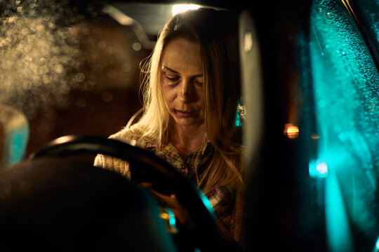 A young Caucasian woman, bearing the painful signs of domestic abuse with visible bruises and blackened eyes, drives her car at night