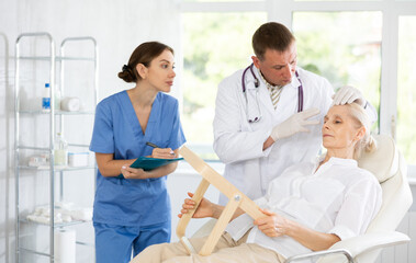 Obraz premium Man dermatologist examining female face skin. Mature woman getting professional help during appointment visit to expert in cosmetology and dermatology, female assistant write recommendations for care