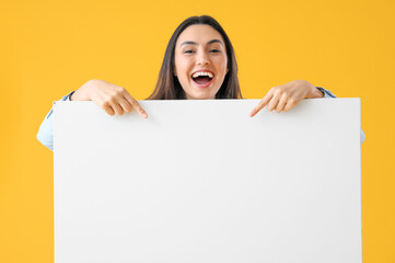 Happy young woman pointing at big blank poster on yellow background